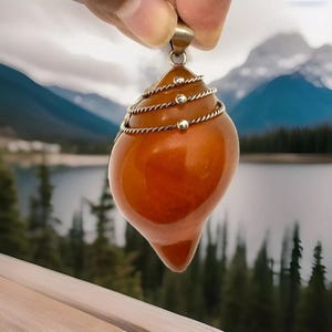 May include: A pendant made of amber with a silver wire wrapped around it. The amber is a deep orange color and has a smooth, polished finish. The pendant is hanging from a silver chain.