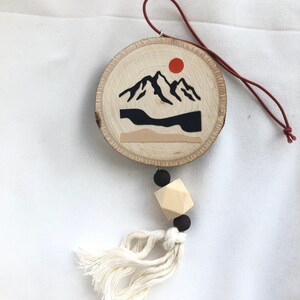 Könnte beinhalten: Ein Holz-Anhänger mit einer bemalten Bergszene und einer roten Sonne. Der Anhänger hat eine braune Lederschnur und eine Holzperle mit einem Quasten.