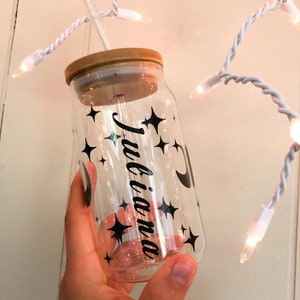 May include: A clear glass reusable water bottle with a bamboo lid and a black straw. The bottle has a black and white design with stars and a crescent moon. The text "Julianna" is written on the bottle.
