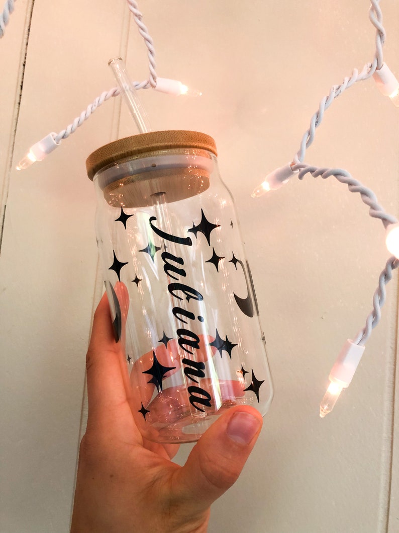 May include: A clear glass reusable water bottle with a bamboo lid and a black straw. The bottle has a black and white design with stars and a crescent moon. The text "Julianna" is written on the bottle.