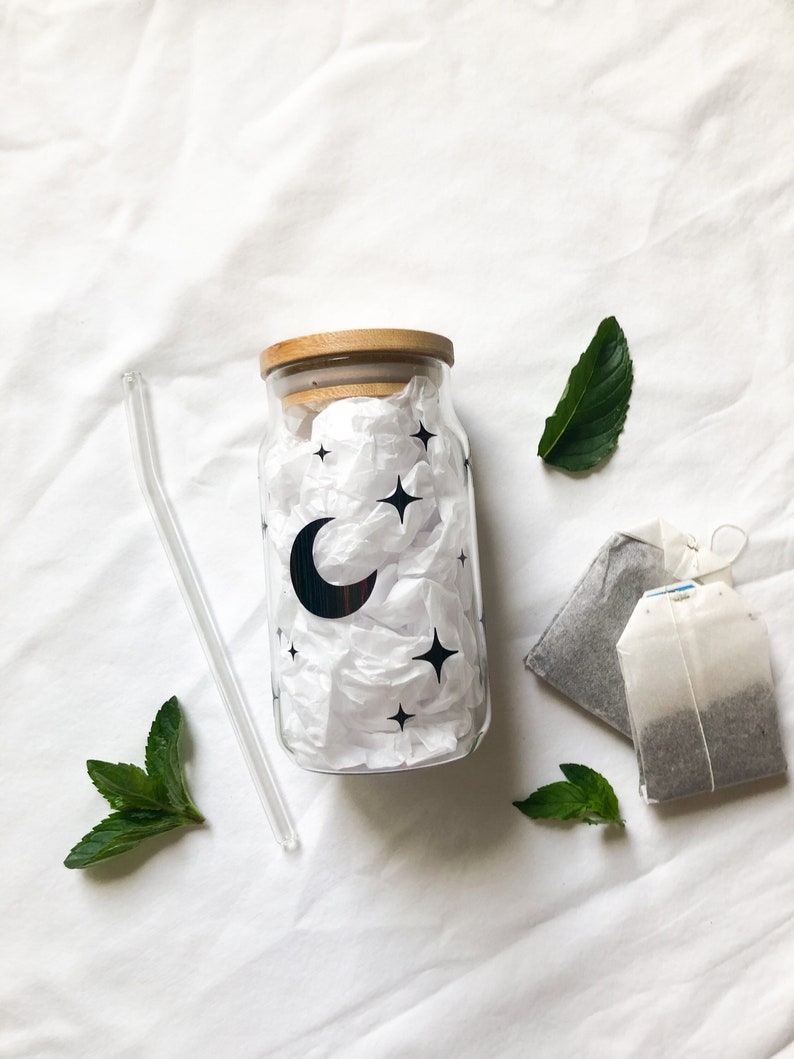 May include: A clear glass jar with a wooden lid and a moon and stars design. The jar is filled with white tissue paper. There are two tea bags and a glass straw next to the jar.