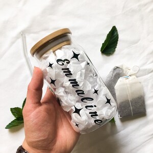 May include: A clear glass bottle with a bamboo lid and a black straw. The bottle is filled with white tissue paper and has black stars and the name "Emmaline" written on it.