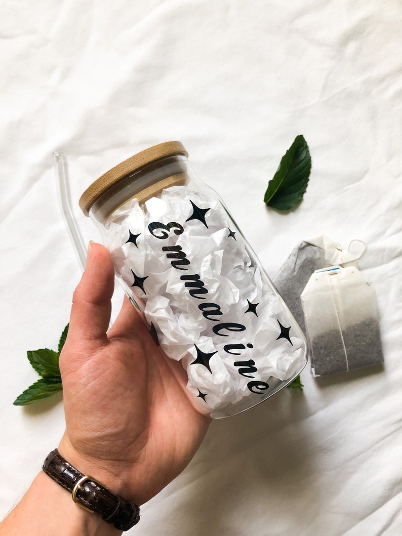 May include: A clear glass bottle with a bamboo lid and a black straw. The bottle is filled with white tissue paper and has black stars and the name "Emmaline" written on it.