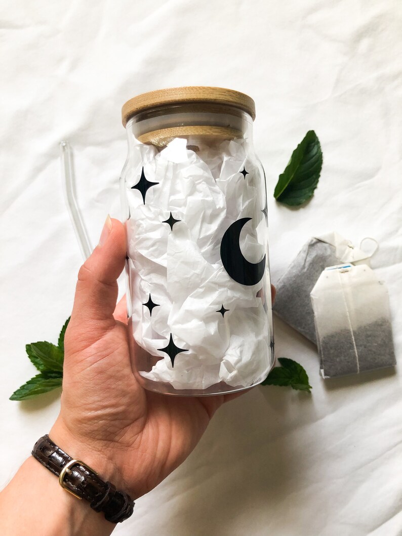 May include: A clear glass bottle with a wooden lid and a moon and stars design. The bottle is filled with white tissue paper.