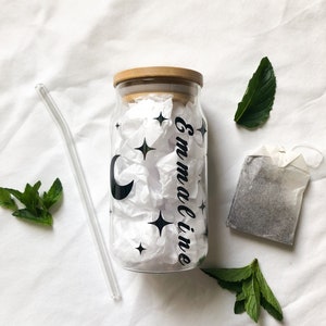 May include: A clear glass bottle with a bamboo lid and a black and white design of a crescent moon and stars. The bottle is labeled "Emmaline". The bottle is accompanied by a glass straw, a tea bag, and fresh mint leaves.