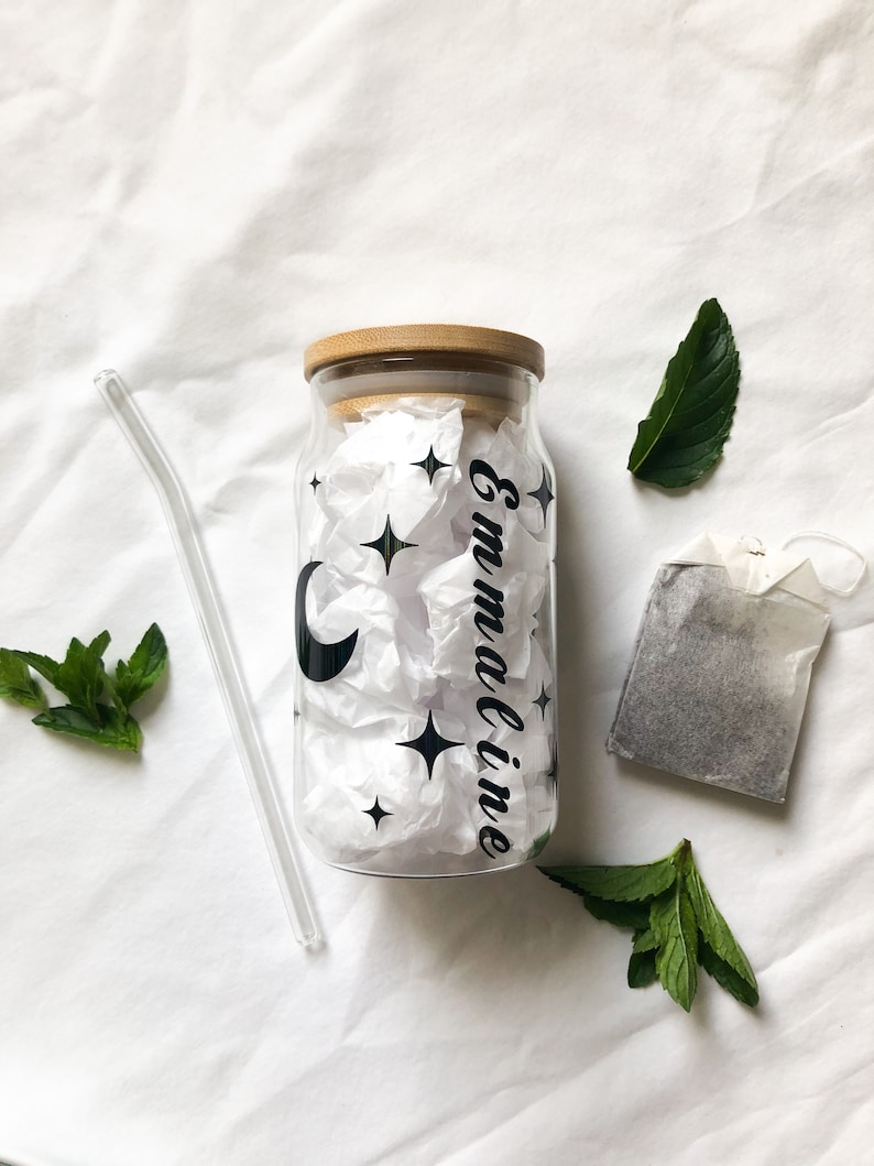 May include: A clear glass bottle with a bamboo lid and a black and white design of a crescent moon and stars. The bottle is labeled "Emmaline". The bottle is accompanied by a glass straw, a tea bag, and fresh mint leaves.