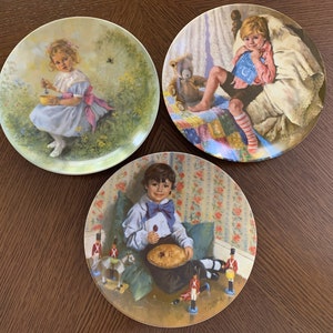 Könnte beinhalten: Drei dekorative Teller mit Kinderillustrationen. Einer zeigt ein Mädchen in blauem Kleid, ein anderer einen Jungen im Bett und der dritte einen Jungen mit einem Kuchen. Die Teller haben eine Vintage-Ästhetik und liegen auf einer Holzoberfläche.