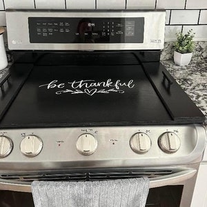 May include: Black stovetop cover with a white inscription that reads "be thankful" with a heart and a vine design. The cover is on top of a stainless steel stove with four knobs.