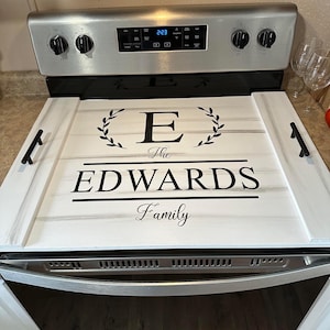 May include: White stovetop cover with a black and white design. The design features a large letter E, the words "The Edwards Family", and a wreath. The cover is made of wood and has two black handles.