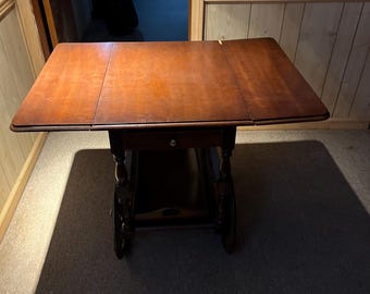 May include: A wooden serving trolley with a drop-leaf top and wheels. The trolley has a drawer and is made of dark wood.