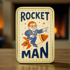 May include: Rectangular tin with the words "ROCKET MAN" and an illustration of a man in a suit and star-shaped glasses riding a rocket. The tin has a cream background with a gold border and is on a wooden surface.