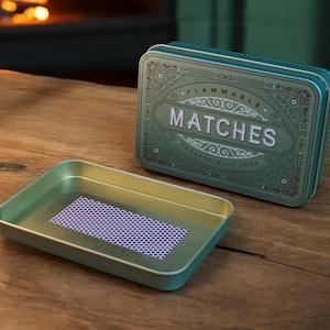 K&ouml;nnte beinhalten: Eine teal-farbene Metall-Streichholzschachtel mit dem Wort &bdquo;MATCHES&ldquo; in wei&szlig;en Buchstaben. Die Schachtel hat ein verziertes Design und ist ge&ouml;ffnet, wodurch ein Tablett mit einer lila und wei&szlig;en gemusterten Schlagfl&auml;che freigelegt wird. Auf einer Holzoberfl&auml;che.