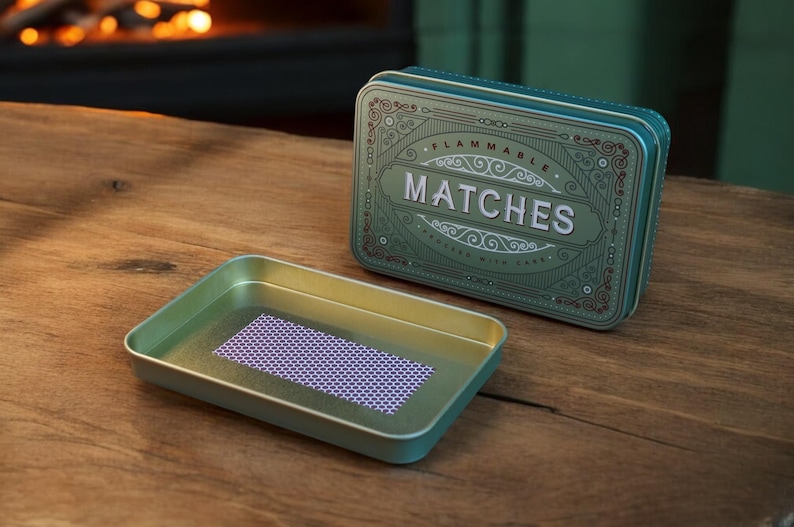 K&ouml;nnte beinhalten: Eine teal-farbene Metall-Streichholzschachtel mit dem Wort &bdquo;MATCHES&ldquo; in wei&szlig;en Buchstaben. Die Schachtel hat ein verziertes Design und ist ge&ouml;ffnet, wodurch ein Tablett mit einer lila und wei&szlig;en gemusterten Schlagfl&auml;che freigelegt wird. Auf einer Holzoberfl&auml;che.