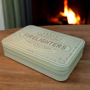 May include: A rectangular, pale green metal tin with the word "FIRE LIGHTERS" in the center. The tin has decorative scrollwork and the words "FLAMMABLE" and "PROCESSED WITH CARE". The tin is on a wooden surface.