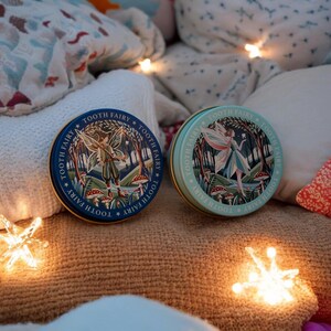 May include: Two round, metal tins with illustrations of fairies in a forest setting. The tins are decorated with the words "Tooth Fairy" repeated around the edges. One tin is blue and the other is green.