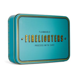 May include: A rectangular, teal metal tin with rounded edges. The lid features gold trim and the words "FLAMMABLE FIRELIGHTERS PROCEED WITH CARE" in gold lettering. The tin is designed to hold firelighters.