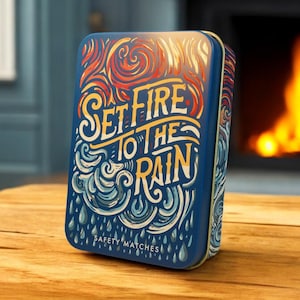 May include: A blue tin with a colourful design featuring flames and waves. The text "Set Fire to the Rain" is written in a stylised font. The bottom of the tin reads "Safety Matches".