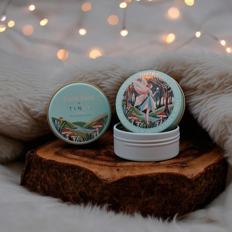 May include: Two round, teal-colored tins with gold lids. The top of the tin on the left says "Tooth Fairy Tin" with a small illustration of a river and forest. The top of the tin on the right says "Tooth Fairy" with a larger illustration of a fairy in a forest.