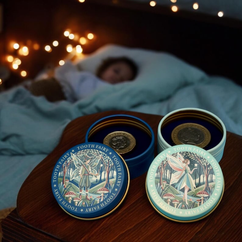 May include: Two small, round, blue and green metal boxes with lids. The lids have a whimsical illustration of a fairy in a forest with mushrooms and the text "*TOOTH FAIRY* TOOTH FAIRY TOOTH FAIRY*" around the edge.