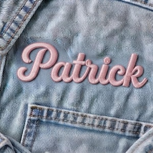 Op de afbeelding: Een lichtblauw acid-wash spijkerjack met de naam Patrick geborduurd in een zachtroze cursief lettertype op de borst. Het kledingstuk is voorzien van zichtbare stiksels, een kraag en een zakflap aan de voorzijde van de denim stof.
