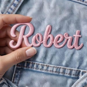 May include: A light pink embroidered patch featuring the name Robert in a cursive script font. The lettering is stitched with a satin thread finish and is positioned against a light blue denim fabric background with visible stitching details.