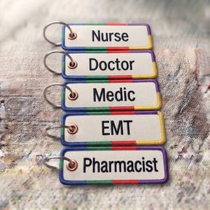 Könnte beinhalten: Fünf rechteckige Schlüsselanhänger aus Stoff mit Metallringen, jeweils mit weißer Mitte und schwarzem Text, der Nurse, Doctor, Medic, EMT und Pharmacist lautet. Die Ränder sind mit abwechselnden blauen, grünen, roten und gelben Segmenten bestickt.