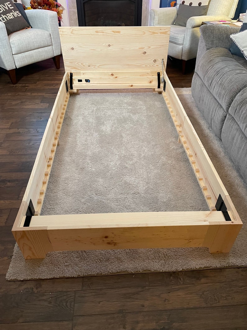 K&ouml;nnte beinhalten: Ein Holzbettgestell mit Lattenrost und einem klappbaren Kopfteil. Der Rahmen ist aus unbehandeltem Kiefernholz gefertigt und hat schwarze Metall Scharniere.