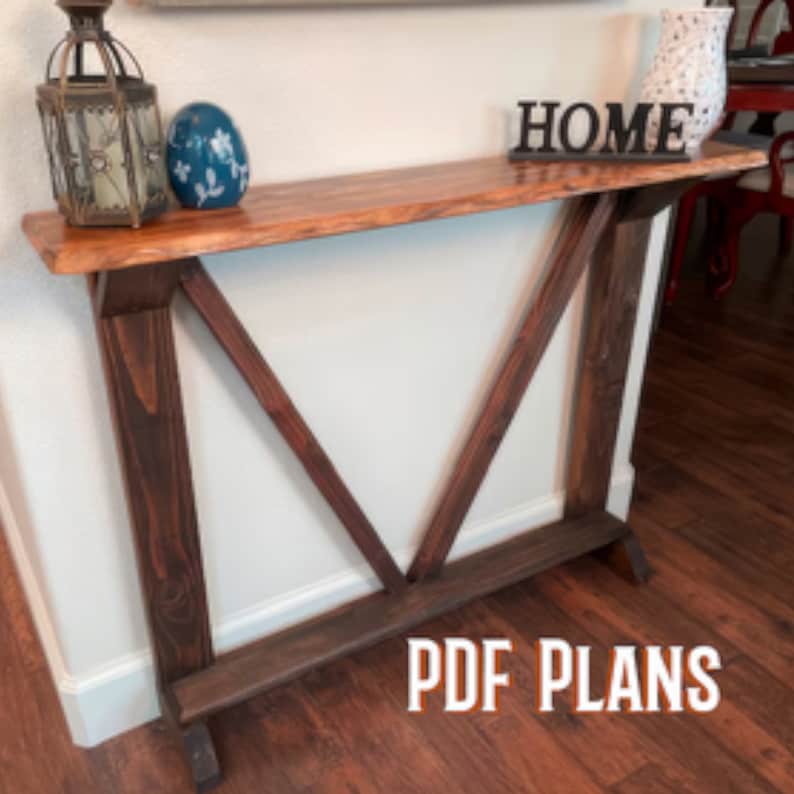 May include: A wooden console table with a brown stained finish and a natural wood top. The table has a simple, modern design with a V-shaped base. The table is decorated with a blue and white ceramic egg, a lantern, and a black wooden sign that says "HOME".