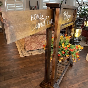 May include: A wooden sign holder with two signs. The top sign says "Welcome" and the bottom sign says "Home Sweet Home". The holder has a shelf with a potted plant and two lanterns.
