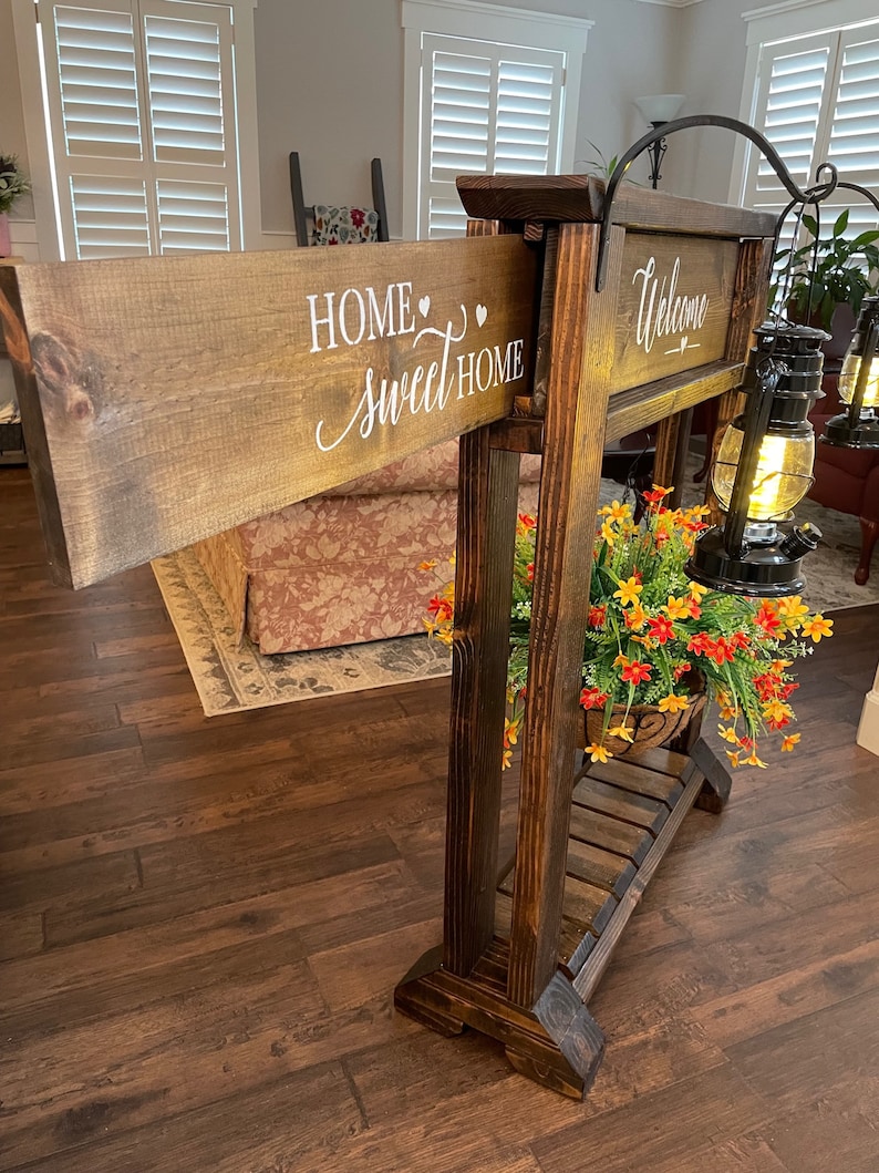 May include: A wooden sign holder with two signs. The top sign says "Welcome" and the bottom sign says "Home Sweet Home". The holder has a shelf with a potted plant and two lanterns.