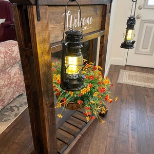 May include: A wooden welcome sign with two black lanterns hanging from hooks. The sign is decorated with a heart and the lanterns are lit. There is a potted plant with orange and yellow flowers hanging below the sign.