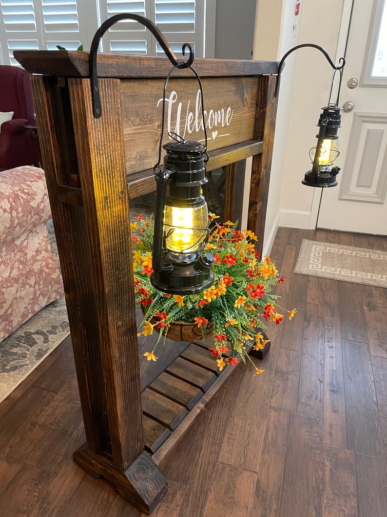 May include: A wooden welcome sign with two black lanterns hanging from hooks. The sign is decorated with a heart and the lanterns are lit. There is a potted plant with orange and yellow flowers hanging below the sign.