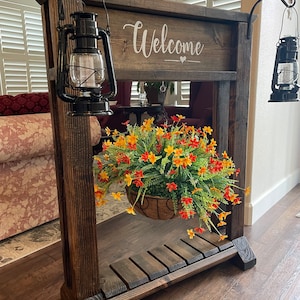May include: A wooden welcome sign with a hanging planter and two black lanterns. The sign says "Welcome" with a heart symbol. The planter has a basket with orange and yellow flowers.