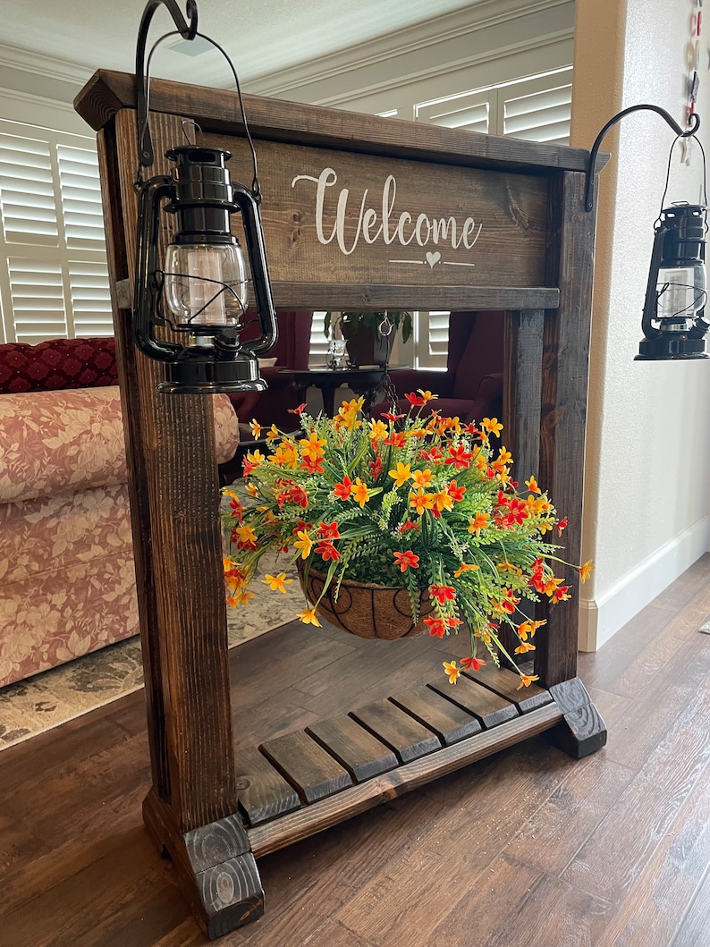 May include: A wooden welcome sign with a hanging planter and two black lanterns. The sign says "Welcome" with a heart symbol. The planter has a basket with orange and yellow flowers.