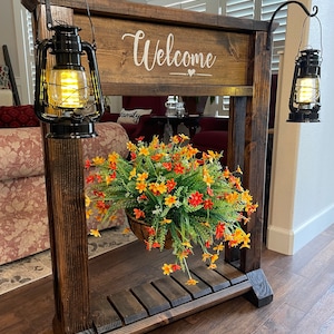 May include: A wooden welcome sign with a hanging planter and two black lanterns. The sign says "Welcome" with a heart symbol. The planter has a basket with green and orange flowers.