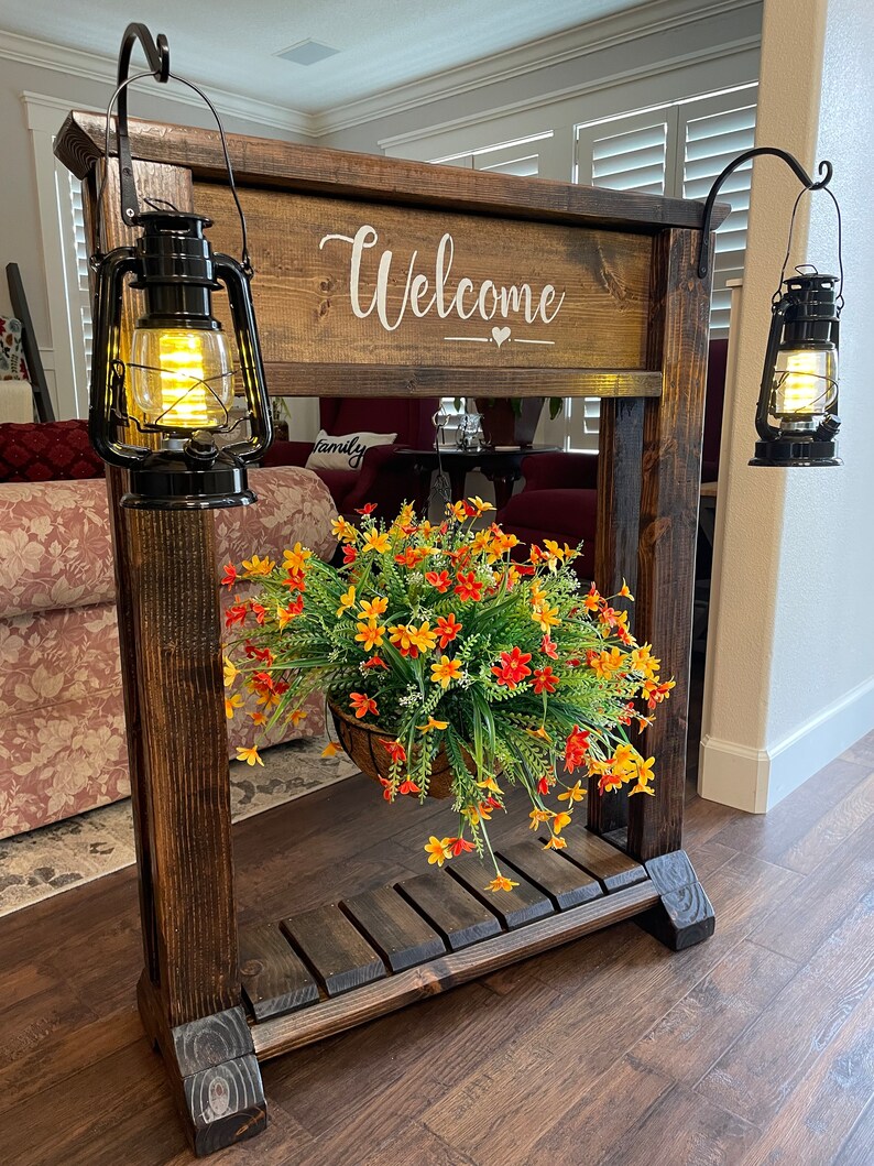 May include: A wooden welcome sign with a hanging planter and two black lanterns. The sign says "Welcome" with a heart symbol. The planter has a basket with green and orange flowers.