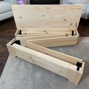 K&ouml;nnte beinhalten: Eine Holzkiste mit einem Scharnierdeckel, ge&ouml;ffnet, um eine Reihe von Holzregalen im Inneren zu zeigen. Die Kiste ist aus unbehandeltem Kiefernholz gefertigt und hat schwarze Metallscharniere.