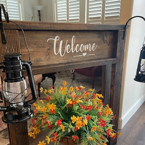May include: A wooden sign with the word "Welcome" painted in white. The sign is hanging on a wall with two black lanterns on either side. A hanging flower basket with yellow and orange flowers is in front of the sign.
