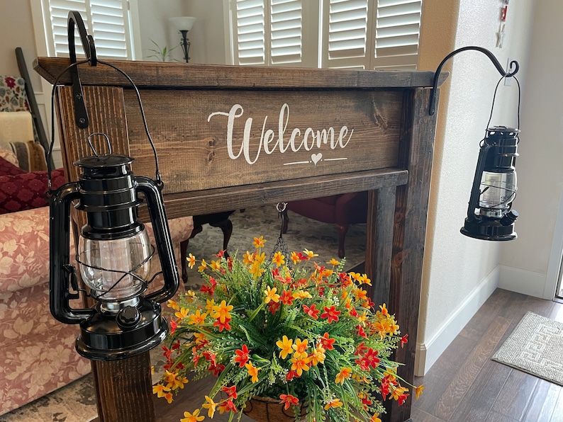 May include: A wooden sign with the word "Welcome" painted in white. The sign is hanging on a wall with two black lanterns on either side. A hanging flower basket with yellow and orange flowers is in front of the sign.