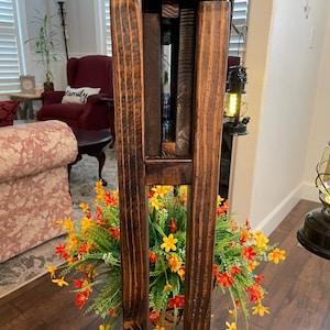 May include: A wooden plant stand with a rustic brown finish. The stand has three shelves and is made of solid wood. The stand is decorated with a potted plant with yellow and orange flowers.