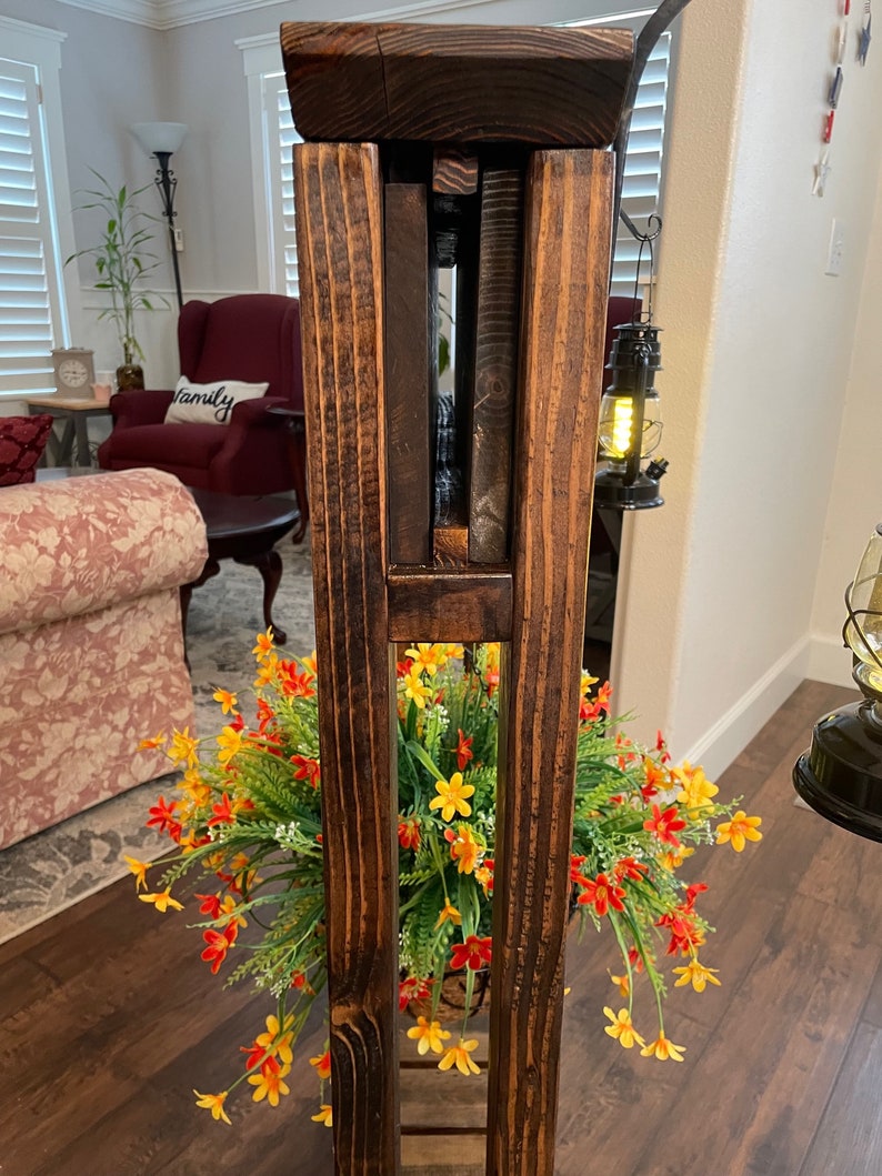 May include: A wooden plant stand with a rustic brown finish. The stand has three shelves and is made of solid wood. The stand is decorated with a potted plant with yellow and orange flowers.