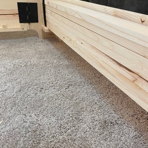 K&ouml;nnte beinhalten: Nahaufnahme eines Holzbettgestells mit einem Lattenrost. Der Rahmen ist aus unbehandeltem Holz und hat ein schwarzes Metallscharnier.