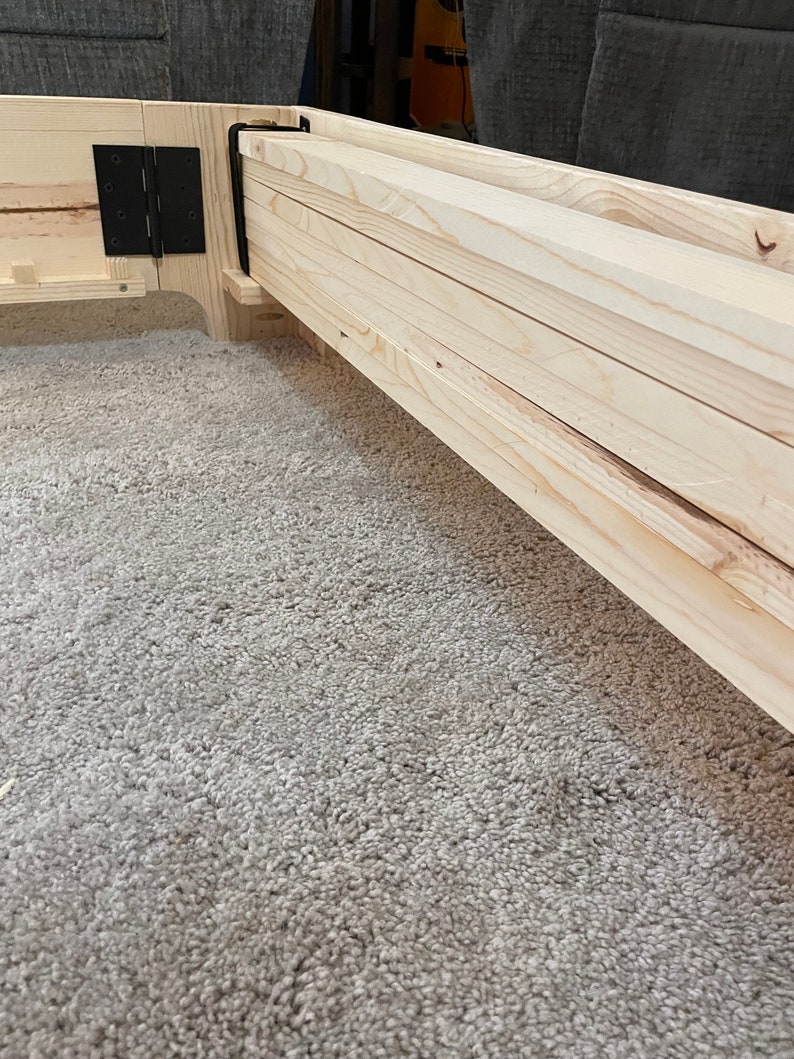K&ouml;nnte beinhalten: Nahaufnahme eines Holzbettgestells mit einem Lattenrost. Der Rahmen ist aus unbehandeltem Holz und hat ein schwarzes Metallscharnier.