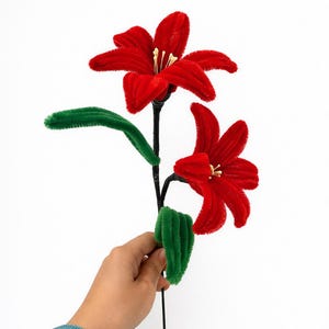 May include: A handcrafted red flower with green leaves and a black stem. The flower has a velvety texture and is set against a white backdrop. The flower's petals are a vibrant red, and the leaves are a bright green.