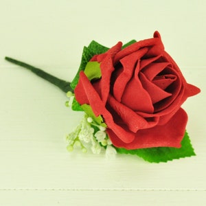 May include: A single, vibrant red rose with detailed petals, green leaves, and a dark green stem. Small white floral accents are clustered near the base of the rose. The artificial flower is set against a white background.