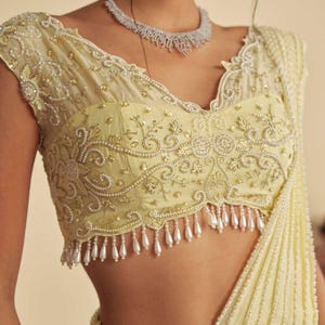 May include: Pale yellow sari with intricate embroidery and pearl accents. The bodice features detailed floral patterns, beadwork, and a scalloped neckline. The sari's fabric is draped elegantly, showcasing the delicate embellishments.