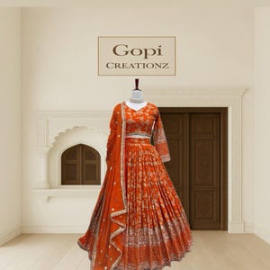 Puede incluir: Un conjunto lehenga choli bordado en naranja y plata sobre un maniquí. El conjunto incluye un top de manga larga, una falda larga y una bufanda a juego. El texto "Gopi Creationz" se muestra encima del maniquí.