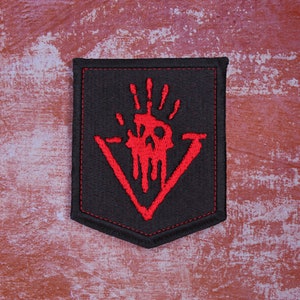 May include: A black embroidered patch featuring a red skull and handprint design. The patch is shaped like a triangle with a point at the bottom.