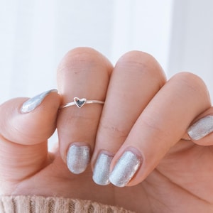 Könnte beinhalten: Ein silberner Ring mit einem kleinen Herzdesign auf der Band. Der Ring wird am Zeigefinger einer Hand mit silbernem Glitzernagellack getragen.