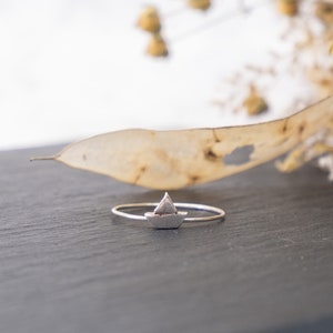 May include: A silver ring with a small sailboat design. The ring is on a dark gray surface.